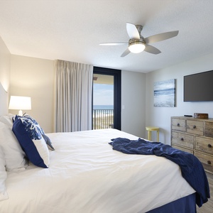 Intimate master bedroom featuring a king bed with decorative pillows, a mounted TV, and a balcony offering views of the beach