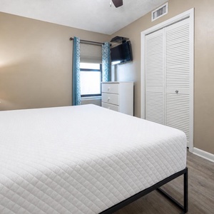 Brightly lit bedroom featuring a comfortable king bed, a dresser, and a TV, with decorative curtains framing the window