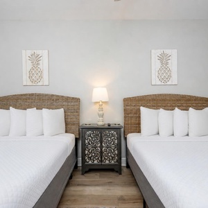 Two queen beds with wicker headboards and white linens are flanked by a decorative nightstand and pineapple-themed wall art