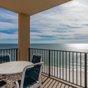 The balcony features a round table with chairs, offering a serene view of the beach and shimmering waters beyond