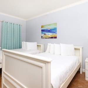 This cozy bedroom features two white beds, soft blue curtains, and a vibrant beach-themed painting for a relaxing stay