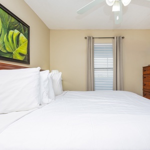 This bedroom features a plush white bed, a wooden dresser, wall-mounted TV, tropical artwork, and natural light
