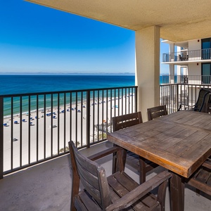 A balcony features a table with six chairs, offering a serene view of the beach and vibrant blue umbrellas below
