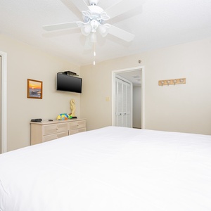 A bright guest bedroom features a king bed, a mounted TV, and a dresser, with a beachy decor accentuating the relaxing vibe