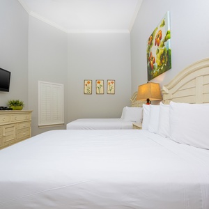 This bright guest bedroom features two queen beds, a mounted TV, and charming floral artwork, creating a welcoming atmosphere