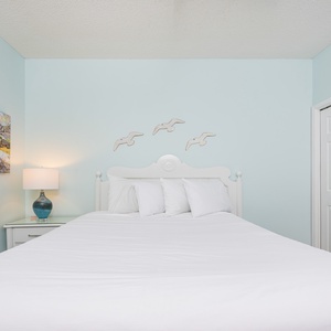 Bright guest bedroom featuring a queen bed, coastal artwork, and a bedside lamp against a soft blue wall