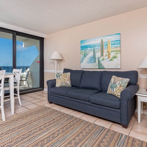 The living room features a comfortable couch, a dining table, and stunning views of the beach through large windows