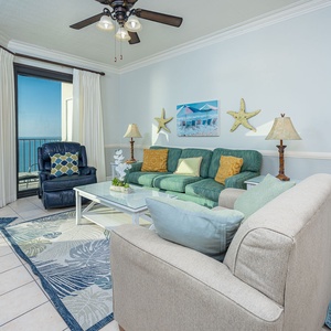 Cozy living area features a green sofa with colorful cushions, a blue recliner, and large windows showcasing the beach front