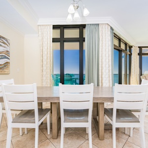This dining area features a light wood table with white chairs, complemented by a stunning beach view through large windows