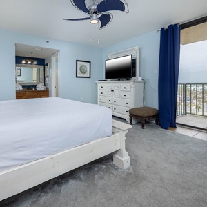 The airy master bedroom features a plush king bed, a TV on a white dresser, and the balcony with a view of the surroundings