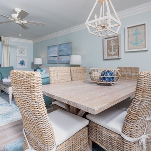The dining area features a rustic wood table surrounded by woven chairs, with beach-themed decor and a view of the beach