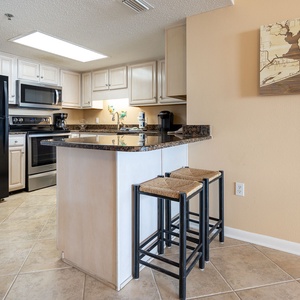 Modern kitchen features dark granite countertops, black appliances, and a breakfast bar with two woven stools