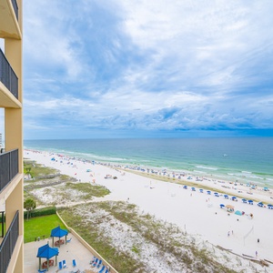 A panoramic view reveals a pristine beach with white sand, dotted with blue umbrellas and gentle waves under a clear sky