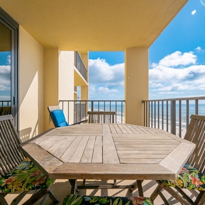A wooden dining table with colorful cushioned chairs overlooks a serene beach front, framed by a clear blue sky