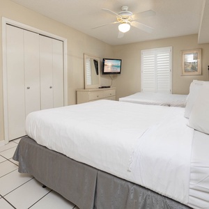 Bright guest bedroom featuring two queen beds, a TV, and a dresser, with natural light filtering through shuttered windows