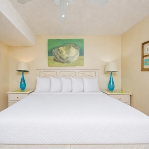 A bright bedroom features a king bed with plush white bedding, complemented by turquoise lamps and coastal artwork