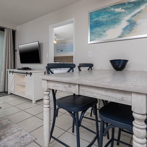 The living area features a large TV, a cozy dining table, and a stunning beachfront view through sliding glass doors