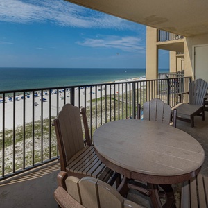 A spacious balcony features a round table and wooden chairs, offering a breathtaking view of the beach and turquoise waters