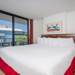 The bedroom features a large bed with crisp linens, vibrant red accents, and a view of the balcony through expansive windows