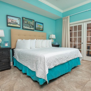 The master bedroom features a cozy bed with white linens, turquoise accents, and French doors leading to a lovely view