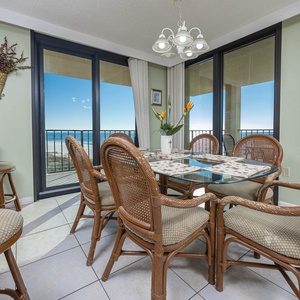 The dining area features a glass-top table surrounded by wicker chairs, with expansive views of the beach