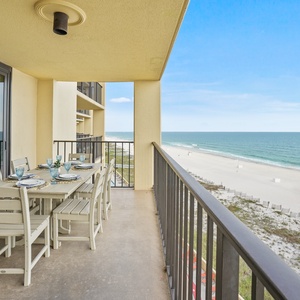 The balcony features a dining table set for six, offering a panoramic view of the tranquil beach and sparkling waters