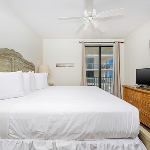 This cozy bedroom features a king bed, a TV, and a sliding door leading to a balcony with a beach view