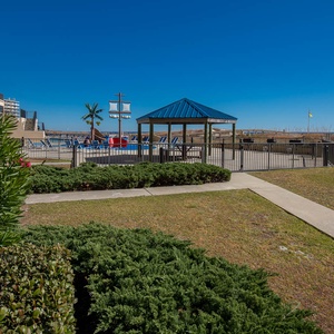 Lush greenery frames the inviting pool area, featuring a shaded gazebo and views of the beach beyond the fence