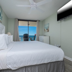Master suite featuring a plush king bed, wall-mounted TV, and a balcony with a view of the beach and seating area