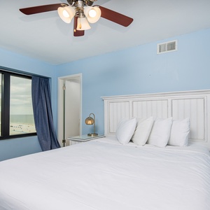This bedroom features a comfortable white bed, blue walls, and a view of the beach through large windows