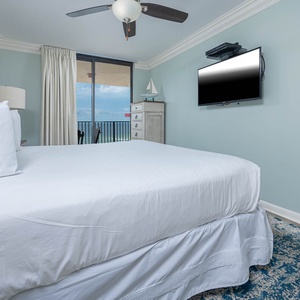 Brightly decorated master bedroom featuring a plush king bed, a mounted TV, and a balcony with a view of the beach