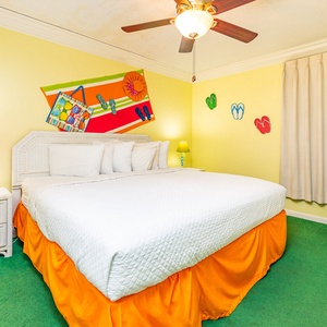 Brightly decorated bedroom featuring a large king bed with white linens, colorful accents, and a TV on a white dresser
