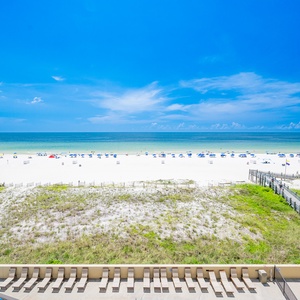 Balcony view showcases white sand and colorful umbrellas lining the beach, framed by clear blue skies and gentle waves