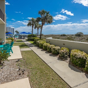 Pathway lined with blooming shrubs leads to the pool area, with views of the beach and gulf visible