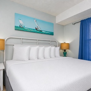 This inviting bedroom features a comfortable bed, stylish lamps, and a vibrant beach-themed artwork for a relaxing stay