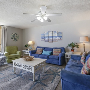 The living room features cozy blue sofas, a light-filled atmosphere, and a serene beach view through the sliding glass door