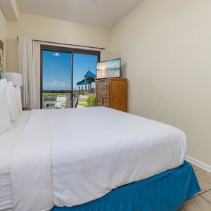 The inviting master bedroom features a plush king bed and a TV, leading to a patio and pool area with a beachfront view