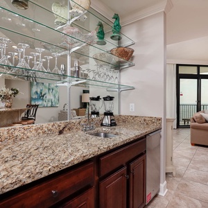 This elegant wet bar features a granite countertop, glassware display, and a view of the living area with large windows