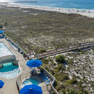 View of the pool area with blue umbrellas and lounge chairs, leading to the beach with white sand and gentle waves