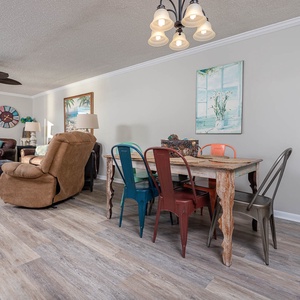 Living area features a mix of colorful chairs around a rustic dining table, with a cozy seating arrangement and beach views