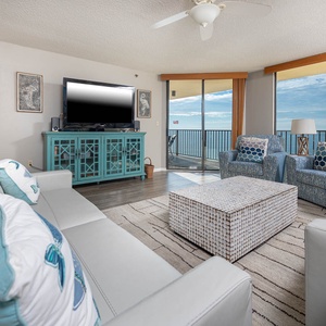 Bright living area features a white leather sofa, blue accent chairs, and a large TV, with a view of the beach beyond