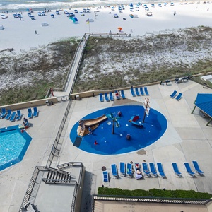 The pool area features a vibrant splash pad and a relaxing pool surrounded by lounge chairs, with the beach just steps away