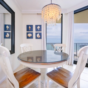 Dining area features a round table with four chairs, framed seashell art, and beachfront balcony