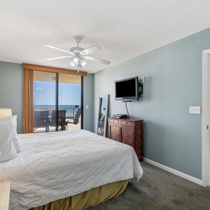 Cozy master bedroom featuring a king bed, a mounted TV, and sliding doors leading to a balcony with a beach front view