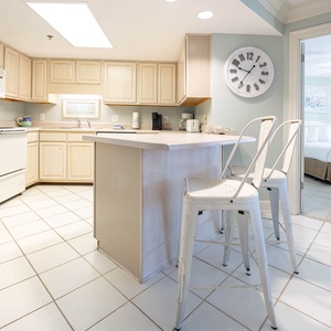 Bright kitchen area features light cabinetry, a breakfast bar with seating, and a view into a cozy master suite beyond