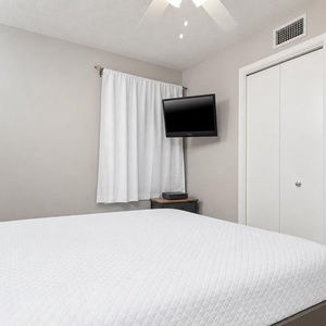 A comfortable bedroom features a large king bed, a wall-mounted TV, and a cozy chair beside a window