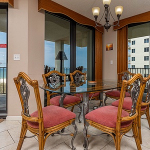 Dining area features a glass table with six cushioned chairs, offering a view of the beach through large sliding doors