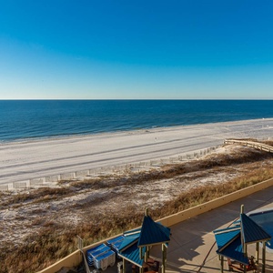 A serene beachfront view showcases white sand and a pool area with shaded seating under blue-roofed gazebos