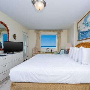 The master bedroom features a large bed, a TV, and a stunning view of the beach through the window