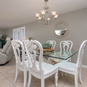 Dining area features a glass table with white chairs, complemented by coastal decor and a view of the balcony